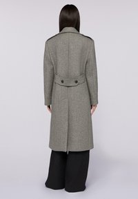 Cappotto di lana grigio con taglio dritto, cintura posteriore, doppio bottone e colletto a punte. Indossato con pantaloni neri larghi.