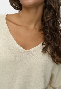 Helle beige gestrickte Pullover mit V-Ausschnitt, locker geschnitten und aus strukturiertem Stoff. Die Nähte sind am Ausschnitt sichtbar.