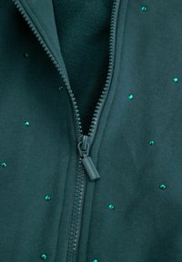 Ciemnoniebieska kurtka zapinana na zamek o gładkiej teksturze, z małymi zielonymi cyrkoniami rozrzuconymi na tkaninie z przodu.