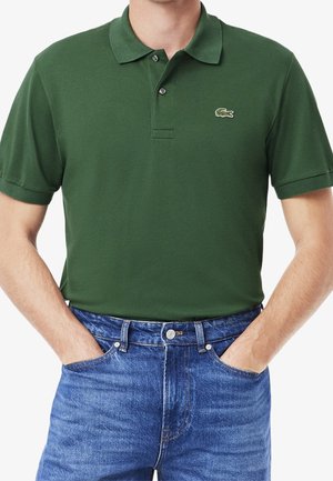 Homme portant un polo manches courtes vert foncé avec un logo crocodile brodé et un jean bleu, les mains dans les poches.