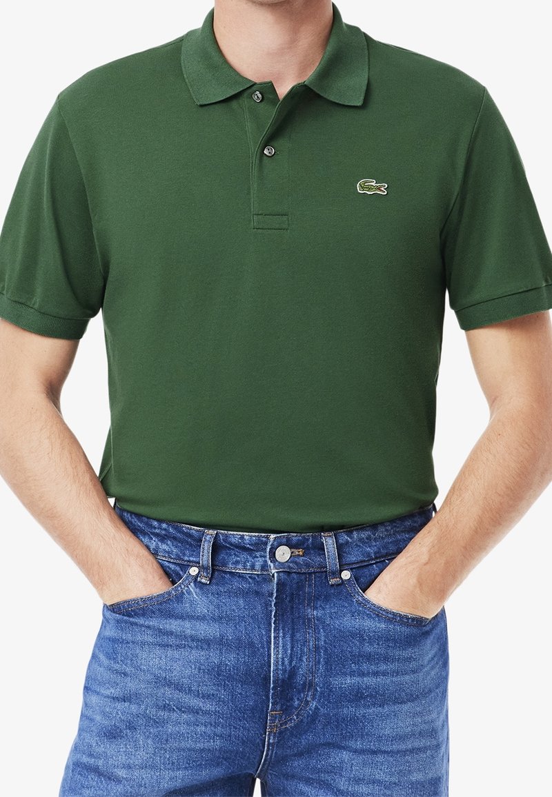 Homme portant un polo manches courtes vert foncé avec un logo crocodile brodé et un jean bleu, les mains dans les poches.