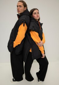 Gilet imbottiti neri con maniche larghe sopra felpe arancioni, abbinati a pantaloni neri larghi. Presentano dettagli con zip e fibbie.