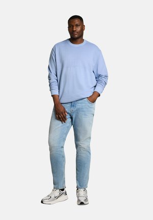 Man standing with one hand in pocket, wearing a light blue sweatshirt, light blue jeans, and white and black sneakers.