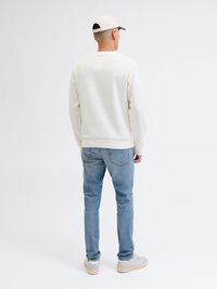 Λευκό πουλόβερ με στρογγυλή λαιμόκοψη, μπλε τζιν και μπεζ αθλητικά παπούτσια. Το καπέλο είναι μπεζ με πράσινο γείσο. Το μοντέλο στέκεται με την πλάτη προς τον θεατή.