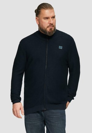 Veste zippée bleu marine foncé avec un col haut, des poignets et un ourlet côtelés, arborant un petit logo carré sur la poitrine gauche. Tissu texturé.