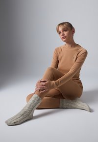 Young woman in beige thermal wear and gray knitted socks sits on floor, holding her knee and looking thoughtfully to the side.