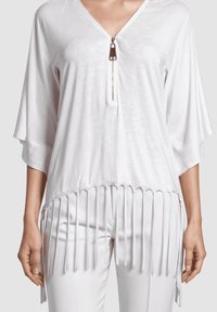 Blusa blanca de cuello en V con cremallera y detalles de flecos largos en el dobladillo, combinada con pantalones blancos a juego.