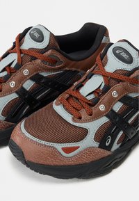 Καφέ και γκρι αθλητικά παπούτσια με δίχτυ και συνθετικά υλικά, μαύρες λεπτομέρειες και πορτοκαλί κορδόνια με ασημένιες οπές για σταθερή εφαρμογή.