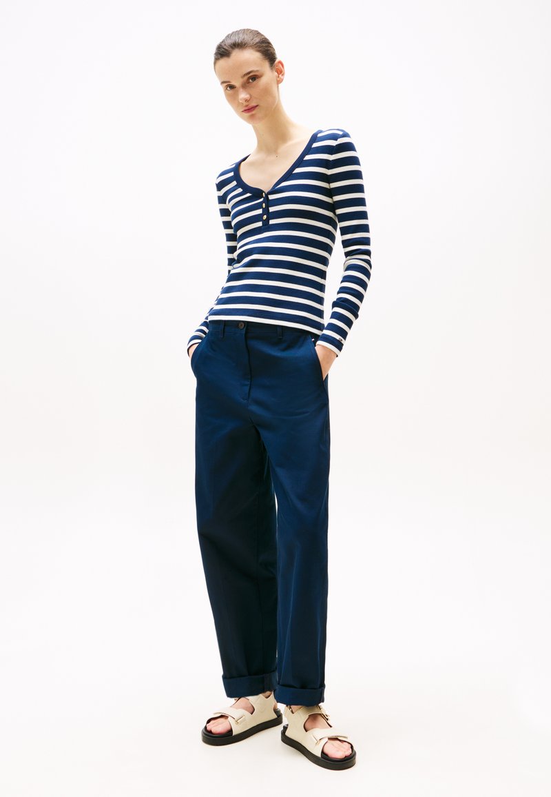 Haut rayé à manches longues en bleu marine et blanc, avec un col en V et des détails de boutons ; assorti à un pantalon bleu marine ample et des sandales crème.