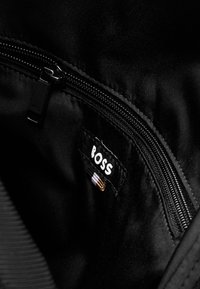 Intérieur d'un sac en tissu noir avec une fermeture éclair, présentant une étiquette blanche "BOSS" et des accents de couture colorés. Texture lisse et design minimal.