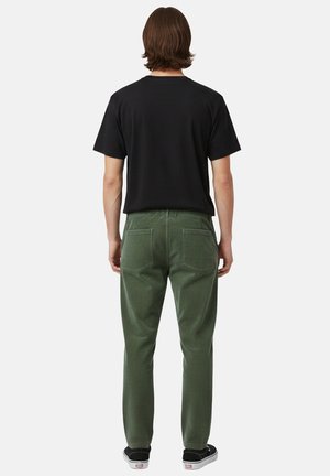 Persona con capelli castani fino alle spalle che indossa una maglietta nera, pantaloni verdi e scarpe da ginnastica nere, in piedi di spalle contro uno sfondo semplice.