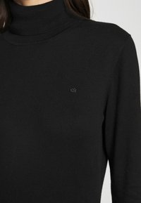 Pull en col roulé noir en tissu tricoté doux. Présente un design ajusté avec un petit logo "ck" brodé sur la poitrine gauche.
