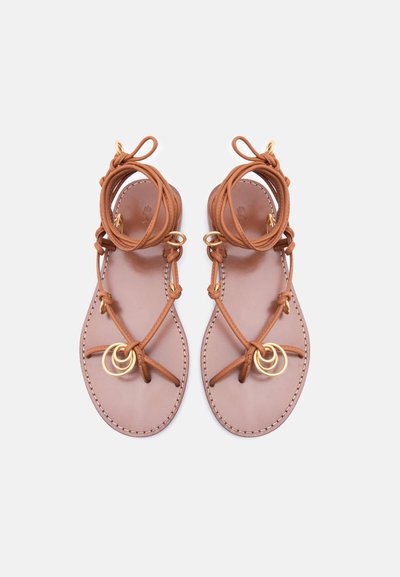 Καφέ δερμάτινα επίπεδα σανδάλια με πολλαπλά λεπτά λουράκια, χρυσές δακτυλίδι διακοσμήσεις και δεσίματα αστραγάλου, σε λευκό φόντο.