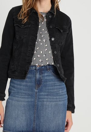Kvinde iført en sort denimjakke, sort-hvidternede bluse med små blomsterprint og en højtaljet blå denimnederdel.
