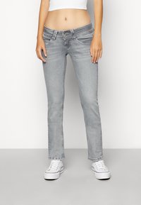 Mujer vestida con jeans de tiro bajo grises claros, una blusa corta blanca y zapatillas de plataforma blancas, de pie contra un fondo sencillo.