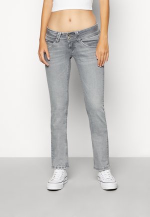 Kvinde iført lysegrå lavtaljede jeans, en hvid cropped top og hvide platform sneakers, stående foran en ensfarvet baggrund.