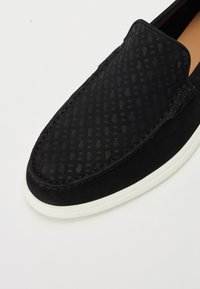 Melni slip-on apavi ar strukturētu augšdaļu, kurai ir austs raksts, un baltu gumijas zoli. Gluda zamšādas materiāla apdare ap papēdi.