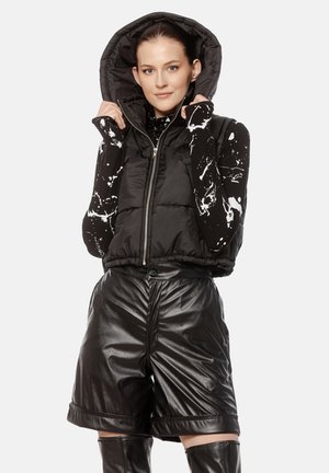 Kobieta ubrana w czarną bezrękawnikową kurtkę puchową z kapturem, czarną z białymi plamami bluzę z długim rękawem, czarne skórzane szorty oraz kozaki do kolan.