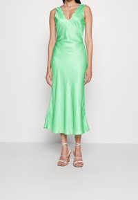 Grünes Satin-Midi-Kleid mit gewelltem Ausschnitt und ausgestellt geschnittenem Saum. Kombiniert mit weißen, mit Riemchen versehenen Absatzsandalen vor einem neutralen Hintergrund.