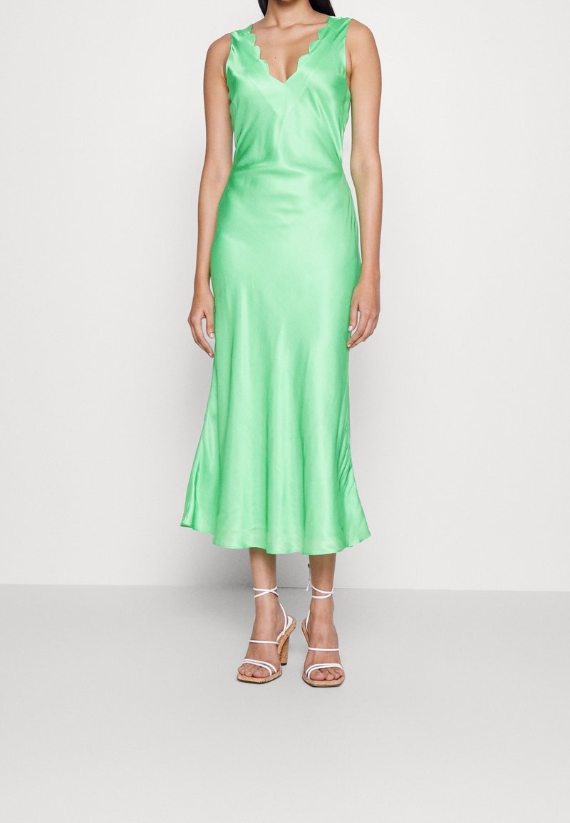 Grünes Satin-Midi-Kleid mit gewelltem Ausschnitt und ausgestellt geschnittenem Saum. Kombiniert mit weißen, mit Riemchen versehenen Absatzsandalen vor einem neutralen Hintergrund.