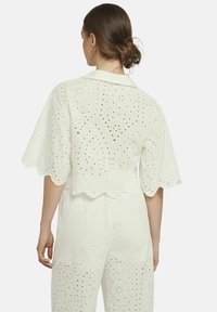Witte katoenen blouse met een gegolfde zoom, korte mouwen en ingewikkelde opengewerkte patronen, gedragen over bijpassende broek.