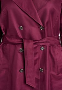 Doppelreihiger Trenchcoat in Burgund mit Taillengürtel, versehen mit glänzenden schwarzen Knöpfen und glattem Stoff mit leichtem Schimmer.