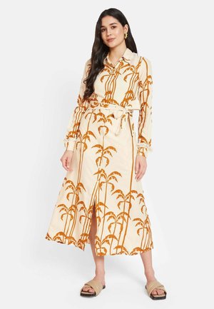 Langarmkleid in Creme mit einem palmbaumartigen Muster in Orange. Mit einem Gürtel, seitlichem Schlitz und gerafften Bündchen. Tragen mit sandfarbenen Rutschpantoletten.