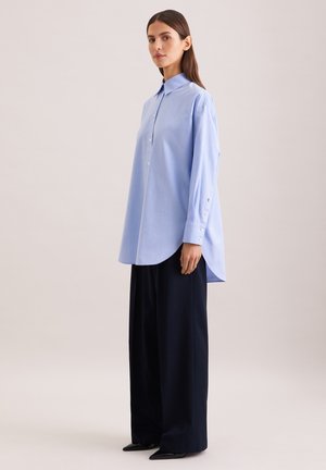 Kvinde iført oversize lyseblå skjorte og vidde sorte bukser, stående foran en ensfarvet beige baggrund.