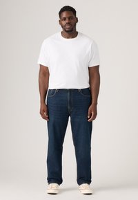 Mann in einem schlichten weißen T-Shirt, dunklen blauen Jeans und weißen Turnschuhen, der vor einem neutralen Hintergrund steht.