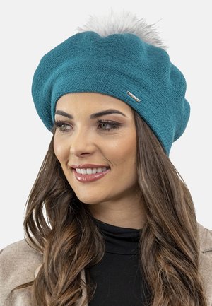 Béret en tricot teal avec un pompon blanc duveteux sur le dessus et un accent de logo métallique. Texture douce, design décontracté, s'adapte bien.