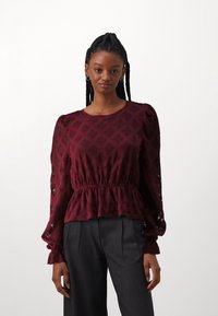 Burgundia peplum-topp tekstuuriga mustriga, pikad varrukad pitsi detailidega ja elastne vöökoht. Kombineeritud hallide kõrge vöökohaga pükstega.