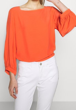 Vrouw die een feloranje blouse met lange mouwen en opgeblazen mouwen draagt, gecombineerd met een wit broek met hoge taille, poserend met één arm omhoog.