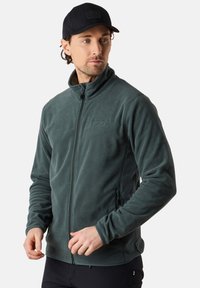Tummanvihreä fleece-takki, jossa on korkea kaulus, vetoketjukiinnitys edessä ja sivutaskut. Takissa on sileä pinta ja hienovarainen logoyksityiskohta.
