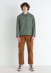Veste zippée verte avec un col, associée à un pantalon marron. Le mannequin porte des baskets dans une combinaison de couleurs blanche et marron.