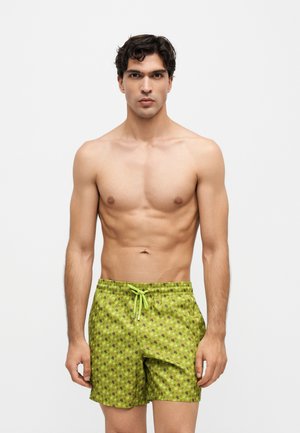 Jeune homme aux cheveux bouclés foncés portant un short de bain vert avec de petits motifs noirs en forme d’étoiles, debout devant un fond clair uni.