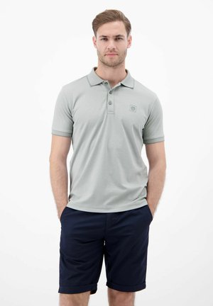 Mężczyzna stojący z rękami w kieszeniach, ubrany w jasno szarą koszulkę polo i ciemnoniebieskie szorty na jednolitym białym tle.