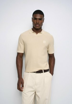 Homme portant un polo beige à manches courtes et un pantalon crème avec une ceinture noire, debout devant un fond clair uni.