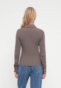 Långärmad, ribbad brun tröja med krage och knäppta manschetter, tillsammans med högmidjade blå denimjeans. Neutral bakgrund.