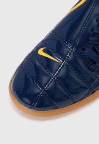 Sapato de verniz azul-marinho com o logo da Nike em amarelo, parte superior texturizada e sola de borracha. Os detalhes incluem painéis cosidos e atacadores.