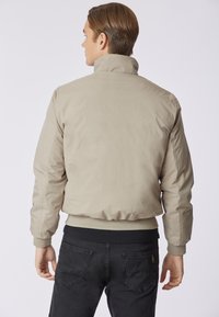 Giacca bomber beige con colletto alto, polsini e vita a coste. Tessuto liscio, chiusura con zip e senza motivi o dettagli visibili.