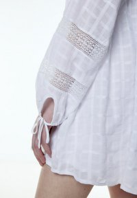 Weißes Kleid mit durchsichtigen Spitzenakzenten an den Ärmeln, strukturiertem Stoff mit Karomuster und einem Band am Handgelenk.