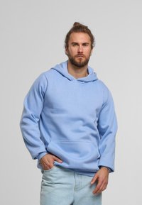 Hoodie di colore azzurro chiaro con tasca frontale, realizzato in tessuto morbido, caratterizzato da una vestibilità comoda e cappuccio con coulisse. Indossato sopra jeans chiari.