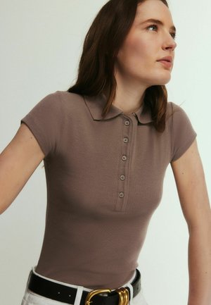 Donna che indossa una camicia taupe a maniche corte con colletto e bottoni, infilata in pantaloni bianchi con cintura nera e fibbia dorata.