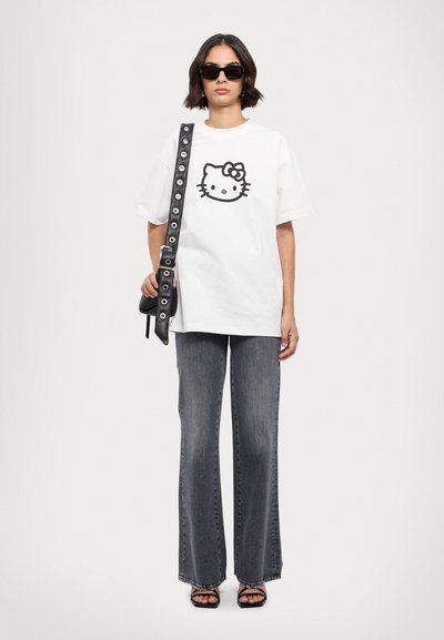 Un tricou alb din bumbac cu un grafic negru Hello Kitty, asortat cu blugi gri din denim cu talie înaltă și evazat și o geantă neagră de umăr.