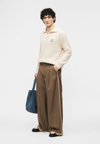 Άνδρας που φοράει κρεμ πουλοβεράκι, φαρδιά καφέ παντελόνια με πλαϊνές ρίγες, μαύρα παπούτσια, κρατώντας μπλε τσαντάκι ώμου, στέκεται μπροστά σε λευκό φόντο.