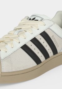 Άσπρο και μπεζ αθλητικό παπούτσι Adidas Superstar με μαύρες ρίγες, κρεμ κορδόνια, υφασμένη σόλα και λογότυπο ορατό στη γλώσσα και την πλευρά.