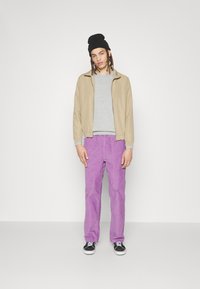 Giacca leggera beige, camicia grigia a maniche lunghe, pantaloni in velluto a coste viola e berretto nero, abbinati a sneakers in bianco e nero.
