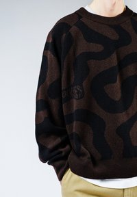 Sweatshirt marron avec un motif ondulé noir, col et poignets côtelés, avec une coupe décontractée. Le matériau semble doux et texturé.