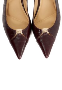 Dark burgundy pointed-toe flats with textured leather and gold-tone metal logo detail on the vamp, viewed from above.