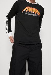 Person som bär en svart långärmad Adidas-tröja med vita ränder på ärmarna och en orange logotyp, ihop med svarta cargobyxor.